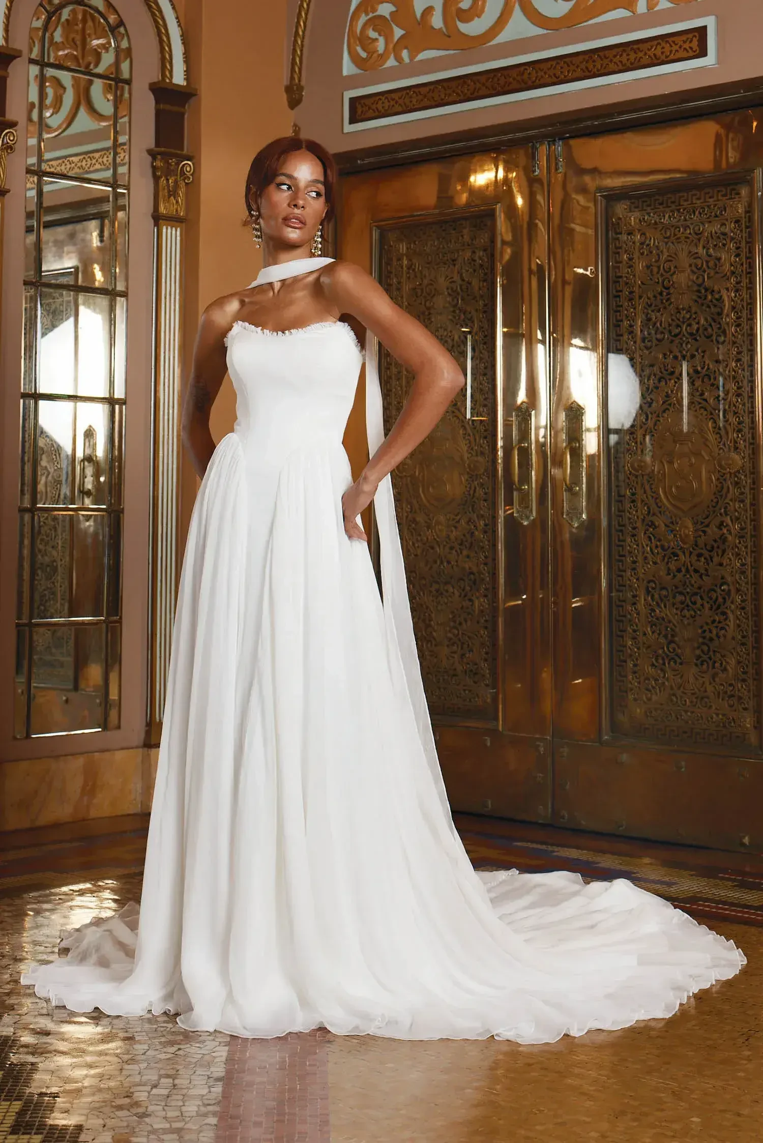 A model wearing a sleek, white wedding gown stands elegantly in a lavish setting with ornate golden doors and intricate decor.