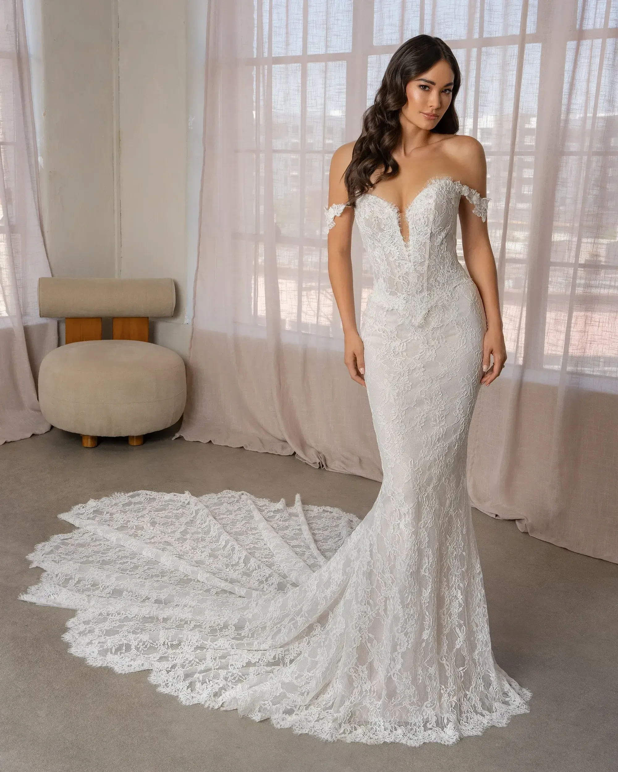 A woman in an elegant off-the-shoulder white lace wedding dress standing in front of a large window, showcasing a long train.